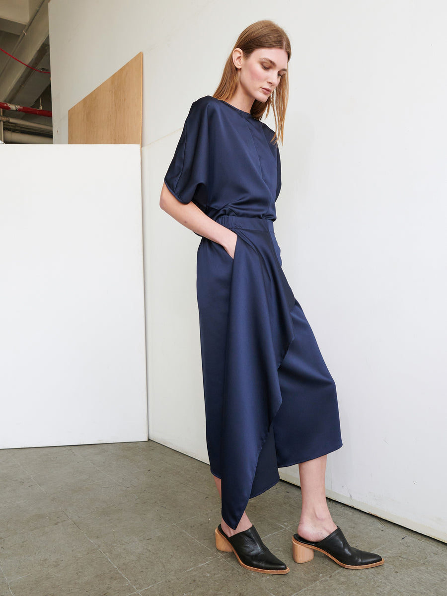 A woman with long brown hair is standing against a white wall in a minimalist indoor setting. She is wearing the One Skirt by Zero + Maria Cornejo, paired with black slip-on shoes that have low heels. One hand is in her pocket, and she appears relaxed.