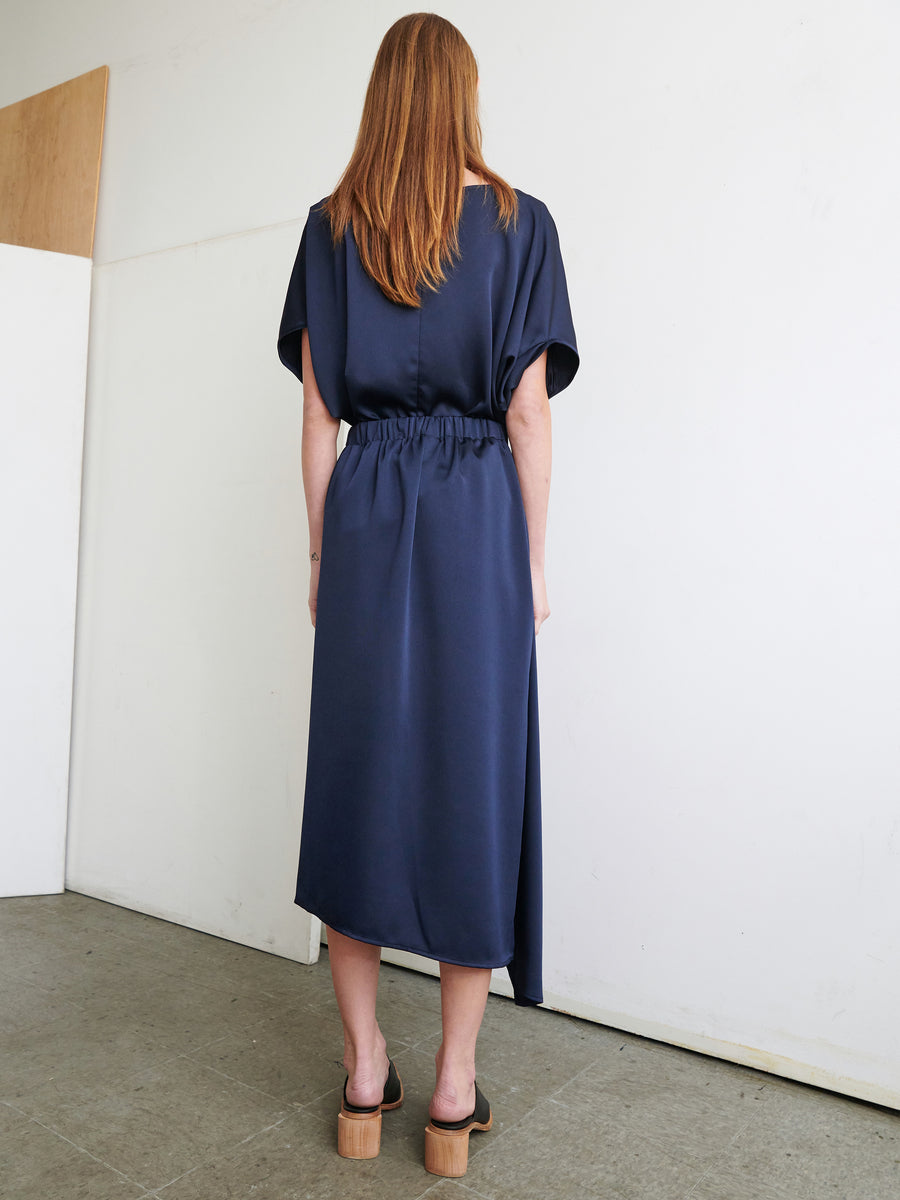 A person with long, light brown hair stands facing a white wall, wearing a navy blue, short-sleeved belted midi dress featuring an asymmetrical front ruffle. They are also sporting black platform sandals with wooden heels. The setting appears to be a minimalistic room. The dress is the 