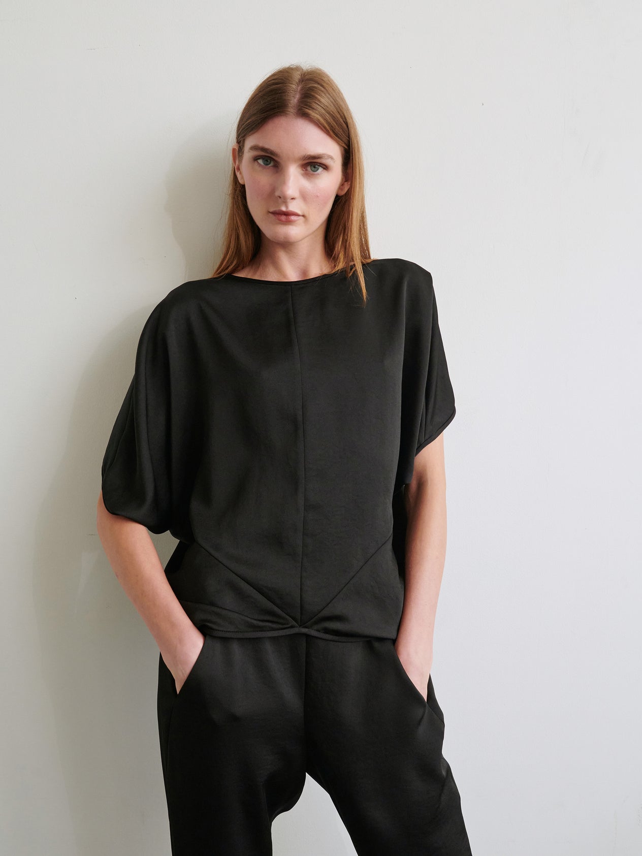 A person with straight, shoulder-length hair is wearing the loose-fitting Rio Top by Zero + Maria Cornejo in dark blue satin. They are standing against a plain off-white background with a neutral expression, and their hands are tucked behind their back.