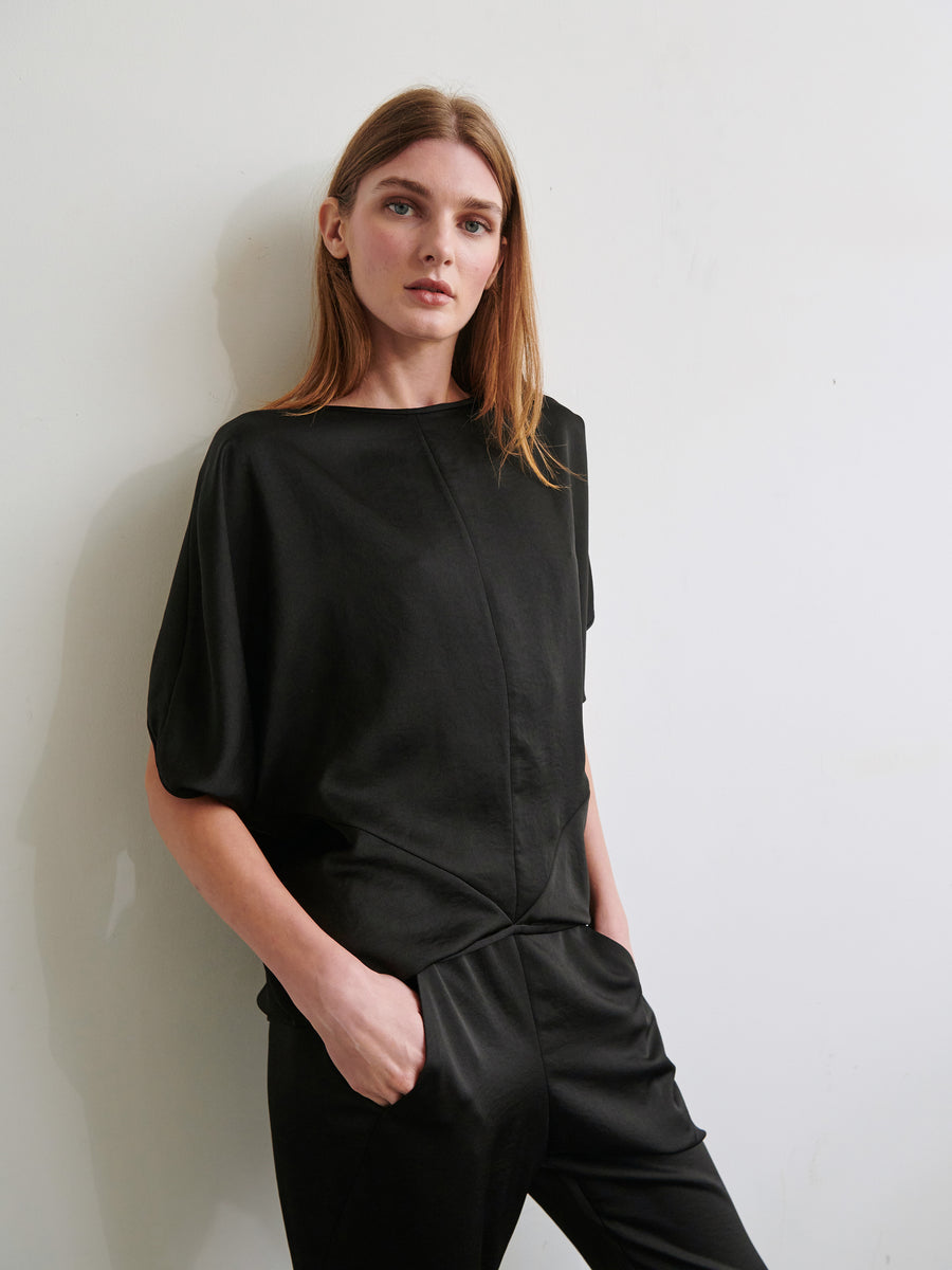 A person with long, light brown hair is wearing a loose-fitting, short-sleeve Rio Top by Zero + Maria Cornejo and black pants. They are leaning against a light-colored wall with one hand in their pocket, looking directly at the camera with a neutral expression.