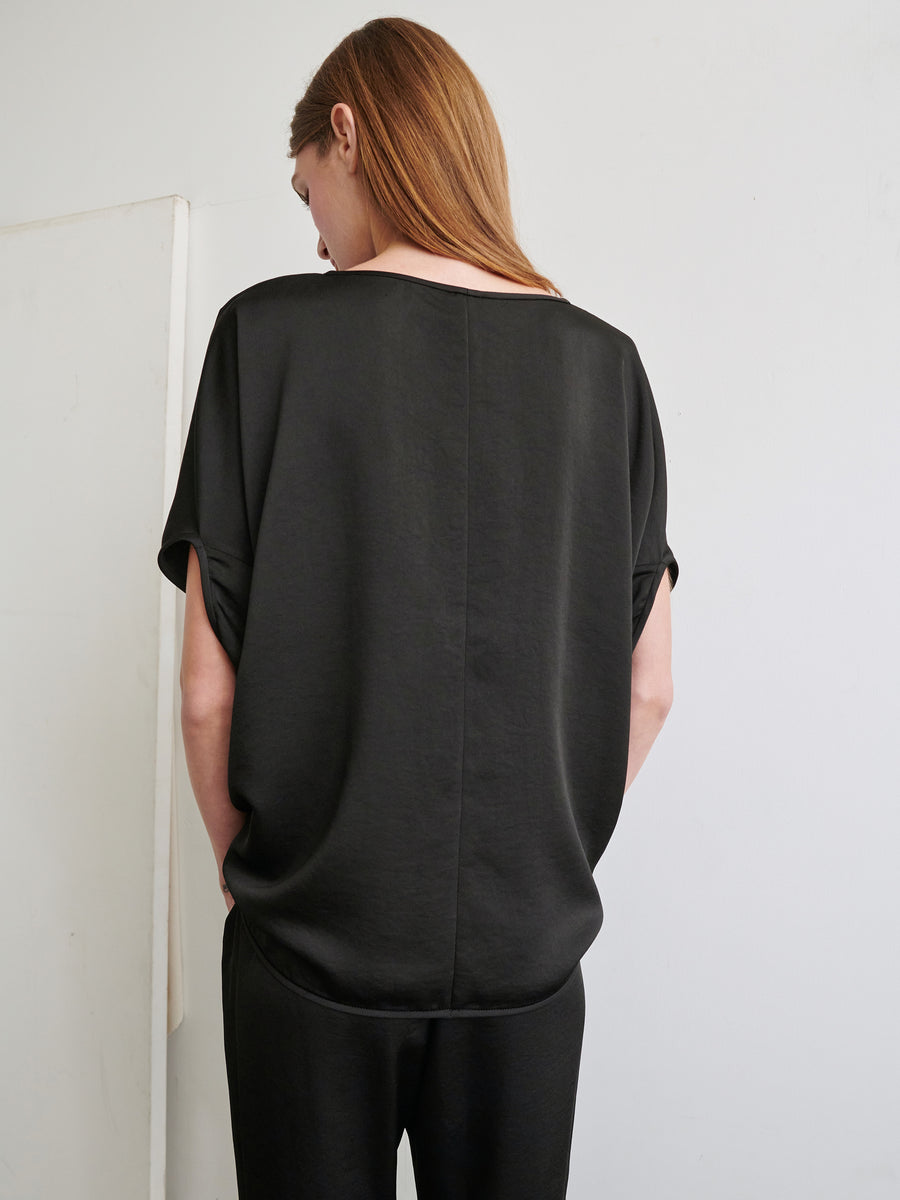 A person with long hair is standing with their back to the camera, wearing the Rio Top, a loose-fitting black shirt made from recycled fabric by Zero + Maria Cornejo, which has rolled-up short sleeves, and black pants. The background is plain and minimalistic, with a light-colored wall.