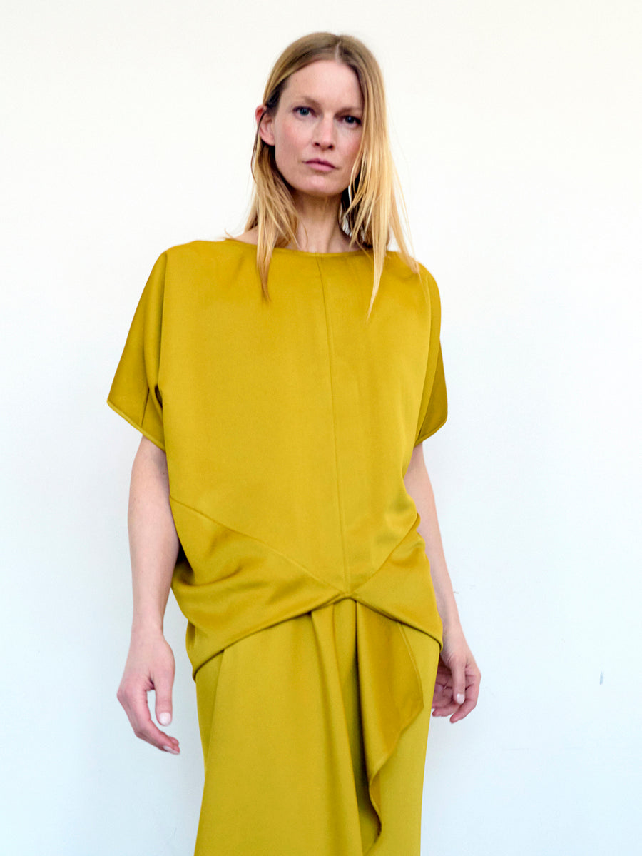 A woman with blonde hair is wearing the Rio Top from Zero + Maria Cornejo, a mustard yellow, short-sleeved recycled drape top featuring an asymmetrical hem, paired with a matching skirt made from 100% recycled fabric. She is standing against a plain white background, looking directly at the camera with a neutral expression.