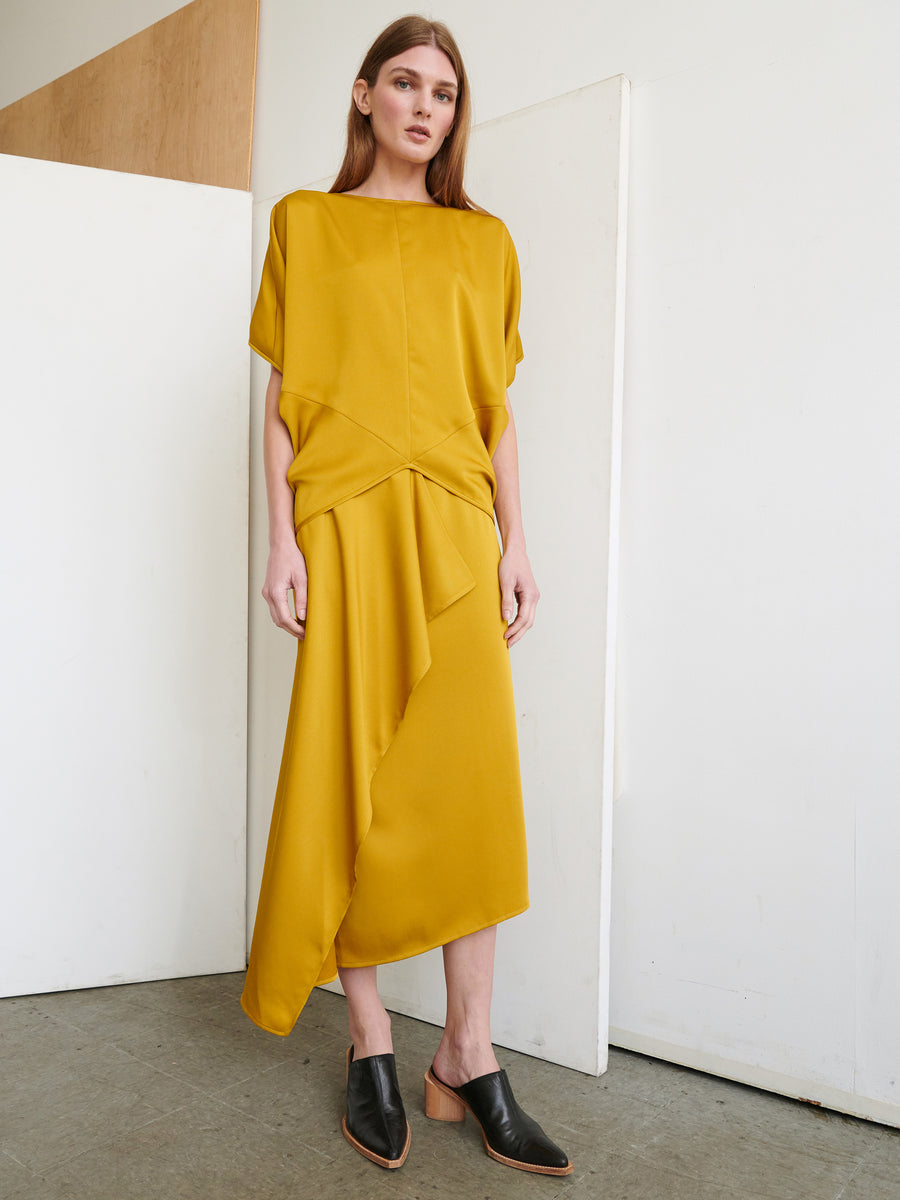 A woman stands in a minimalistic room, showcasing the Rio Top by Zero + Maria Cornejo—a mustard yellow, recycled fabric piece with a unique draped design at the waist. She has long, straight hair and pairs the top with a matching skirt, completing her look with black pointed slip-on shoes. The backdrop features plain white walls and a partial wooden partition.