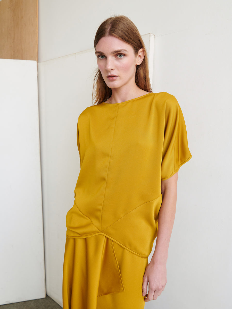 A woman with straight brown hair gazes at the camera, donned in a mustard yellow Rio Top from Zero + Maria Cornejo. The top is crafted from recycled fabric and showcases short sleeves with a simple, elegant design. She poses indoors against a minimal backdrop of white and beige walls, exuding a calm and poised demeanor.