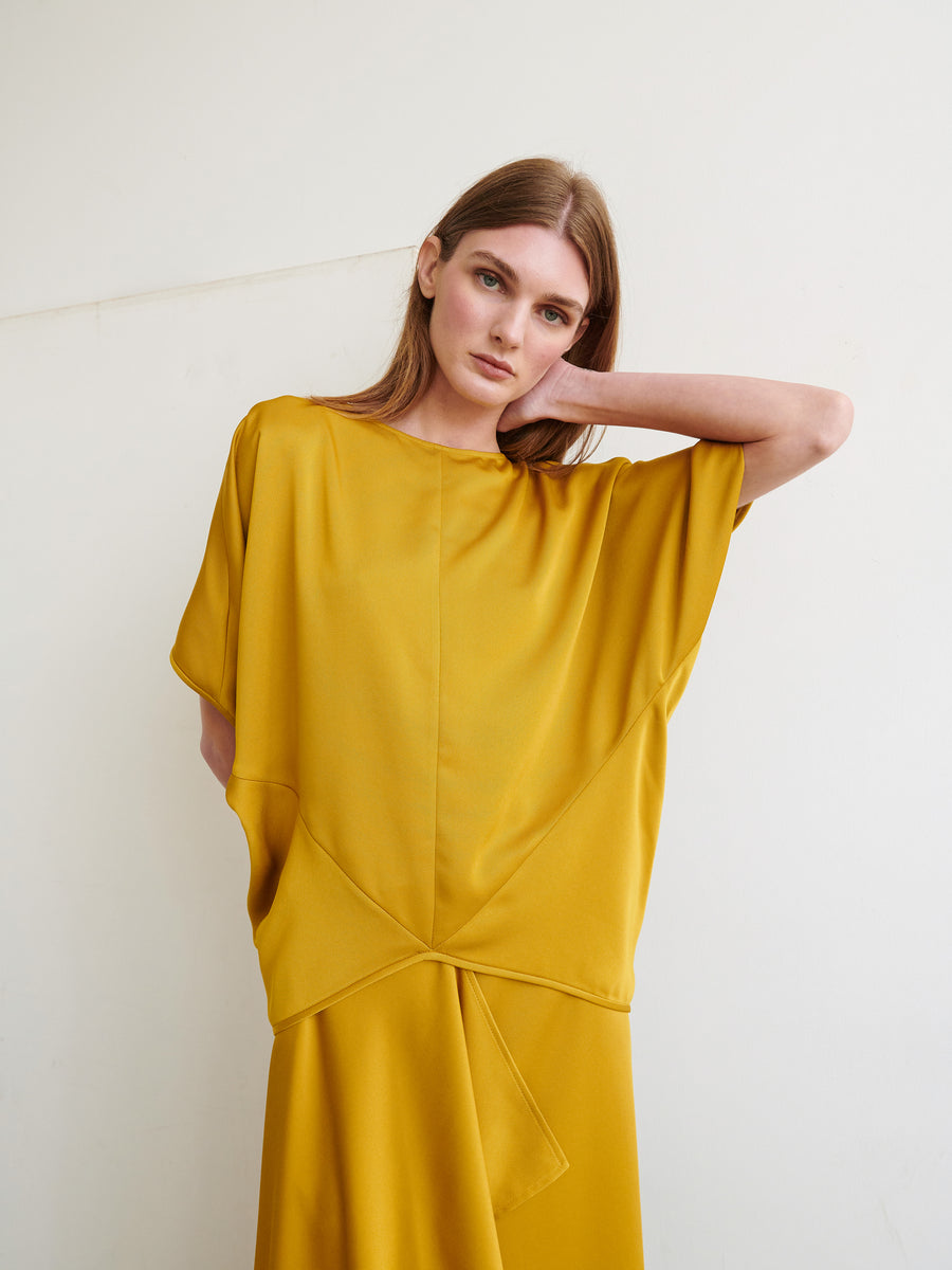 A woman with long, light brown hair is wearing the Rio Top by Zero + Maria Cornejo, made from recycled fabric. She is standing against a plain, light-colored background with one hand resting behind her head and the other in the pocket of her loose-fitting, mustard-yellow dress, gazing directly at the camera.