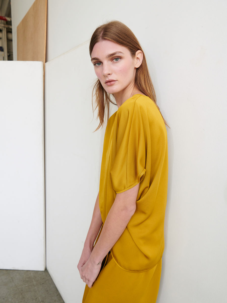 A person with long, brown hair wearing the Rio Top by Zero + Maria Cornejo stands against a white wall. Their gaze is directed slightly away from the camera. The background includes a large white panel and part of a wooden structure.