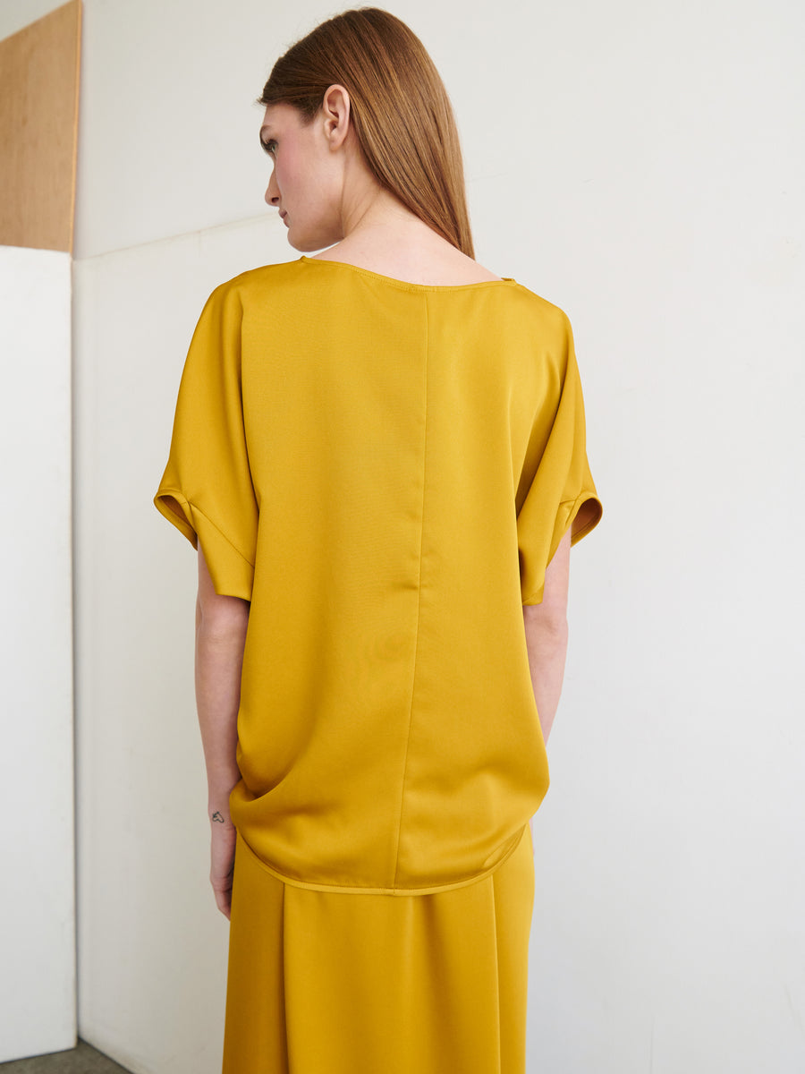 A woman with long, straight hair stands with her back to the camera, wearing the Rio Top and a matching skirt from Zero + Maria Cornejo in a loose-fitting mustard yellow. The outfit is crafted from recycled fabric, features short sleeves and a relaxed fit. Ideal for sustainable fashion, it's easy care: machine wash cold. The background is plain with minimal decor.