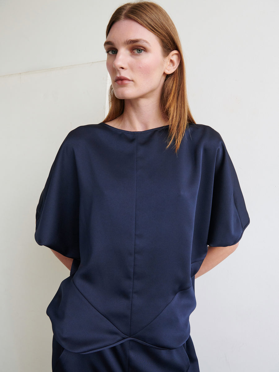 A person with straight, shoulder-length hair is wearing the loose-fitting Rio Top by Zero + Maria Cornejo in dark blue satin. They are standing against a plain off-white background with a neutral expression, and their hands are tucked behind their back.