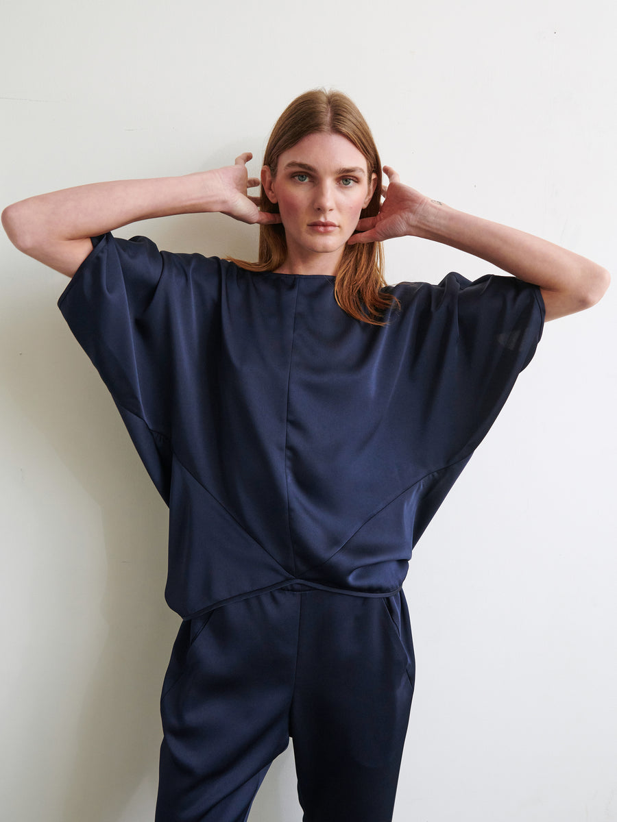 A person with long light brown hair is wearing a loose-fitting, dark blue Rio Top by Zero + Maria Cornejo along with matching pants. They are standing against a plain light-colored background with their hands touching the sides of their head, and their expression is neutral.