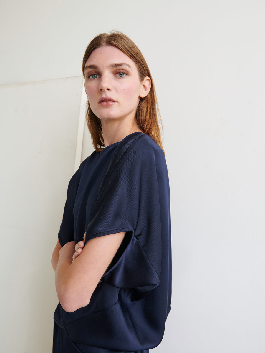 A woman with straight, shoulder-length light brown hair stands against a light-colored wall. She is wearing the navy blue Rio Top from Zero + Maria Cornejo and has her arms crossed. She gazes directly at the camera with a neutral expression.