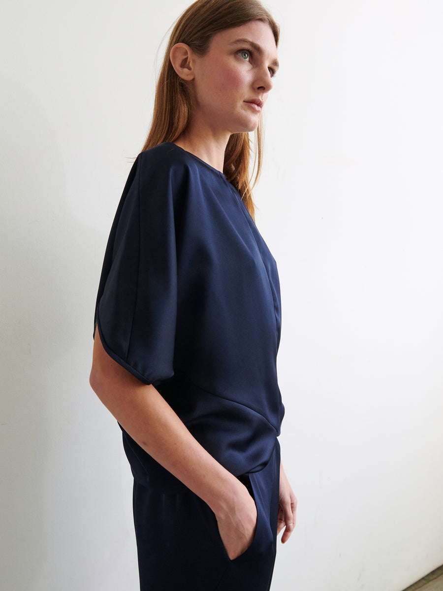 A person with long brown hair is standing in a well-lit room, looking off to the side. They are wearing the loose-fitting, dark blue Rio Top from Zero + Maria Cornejo, which features short sleeves, and have one hand in their pocket. The background is plain and light-colored.