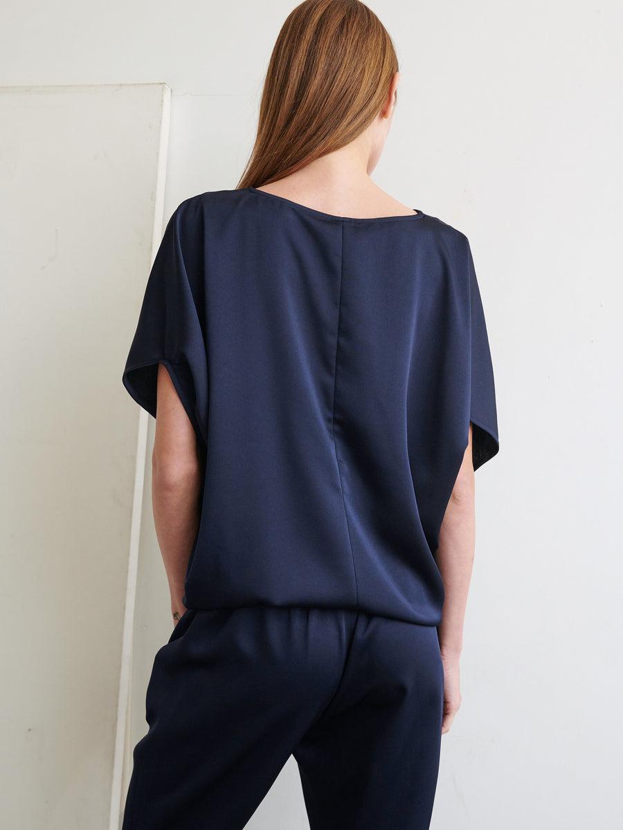 A person with long brown hair is standing with their back to the camera, wearing a loose-fitting, navy blue Rio Top and matching navy trousers made from recycled fabric by Zero + Maria Cornejo. The background is plain with a slight off-white color.