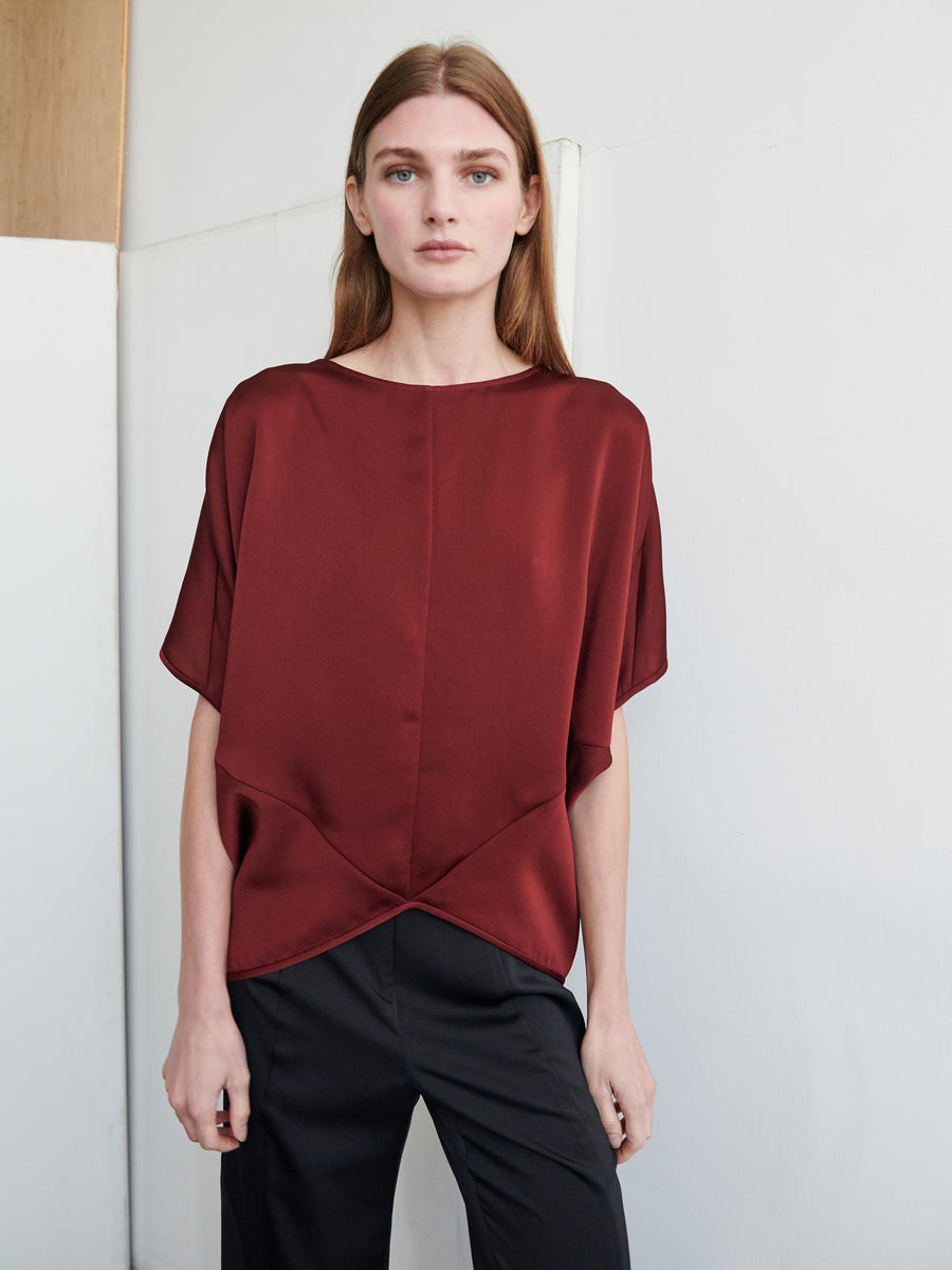 A woman with long brown hair is standing against a simple background, wearing the Zero + Maria Cornejo Rio Top, which is a loose-fitting burgundy recycled drape top with short sleeves and black trousers. The Rio Top, crafted from recycled fabric, features a unique geometric design at the bottom. Her expression is neutral.