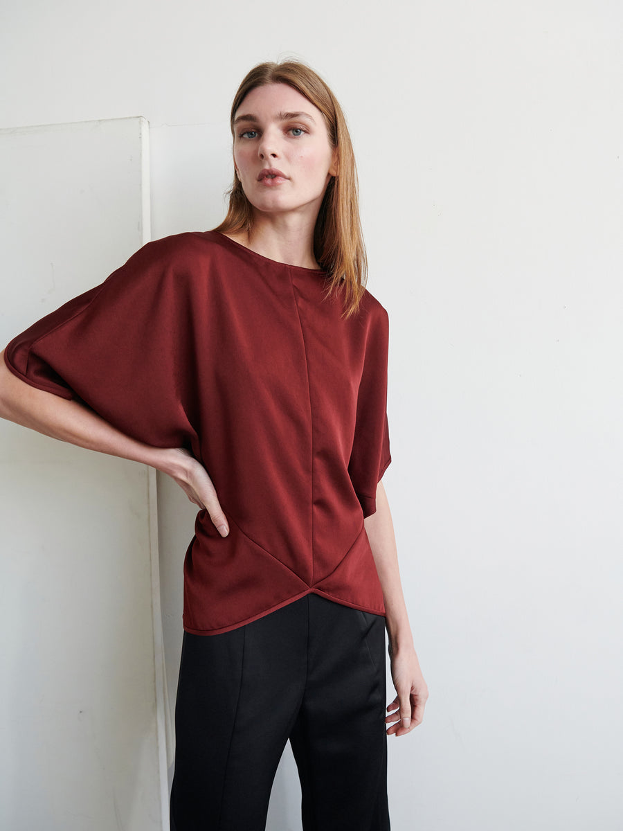 A person with long, light brown hair is wearing the Rio Top in dark red from Zero + Maria Cornejo. The top features a loose fit with dropped shoulders, paired with black pants. They are standing against a plain white background, one hand resting on their hip and a neutral expression on their face.