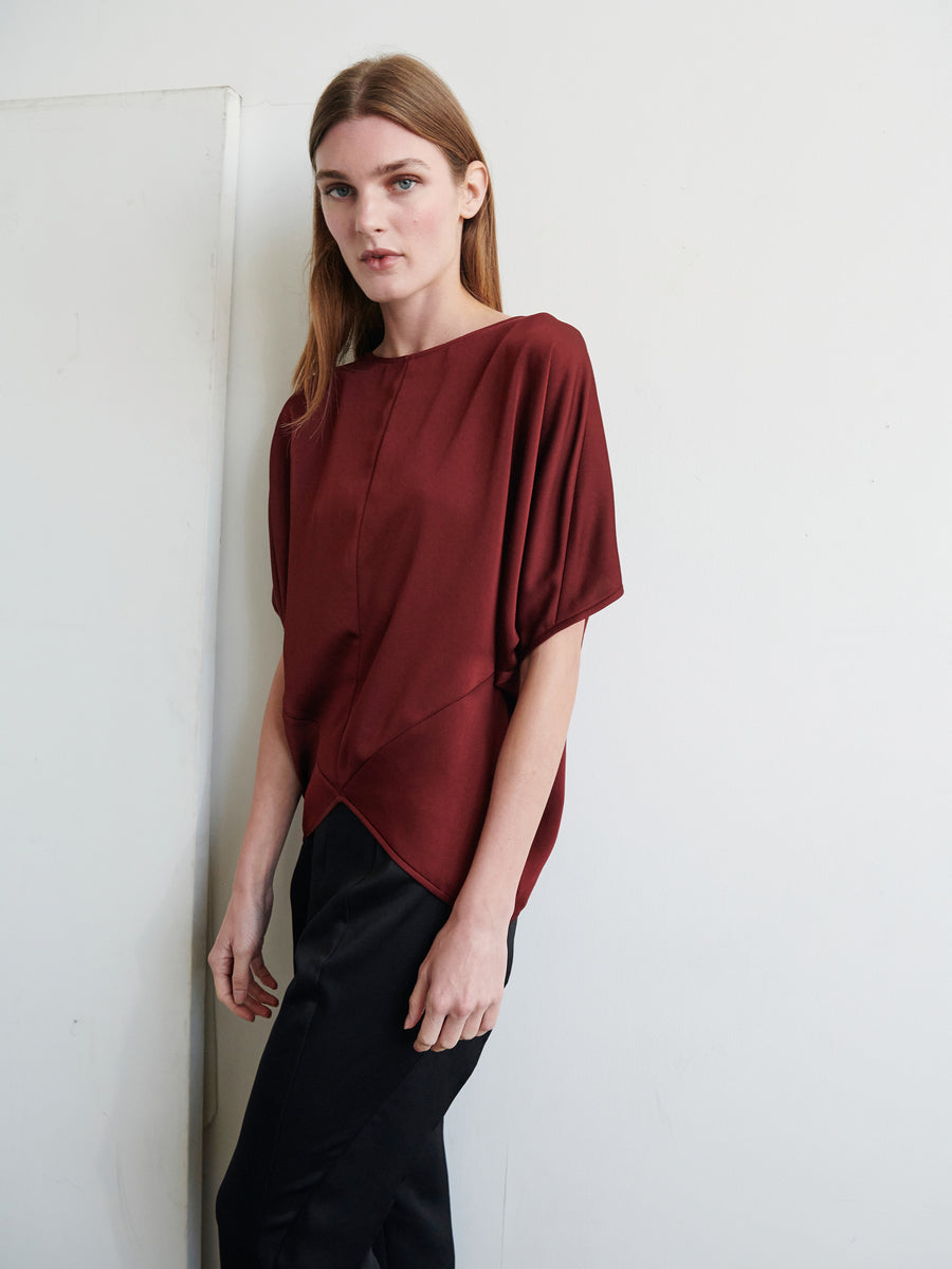 A person with long, straight hair wearing the loose-fitting Rio Top by Zero + Maria Cornejo in dark red, paired with black pants, stands against a white wall, looking at the camera with a neutral expression. The light from the left side casts soft shadows.