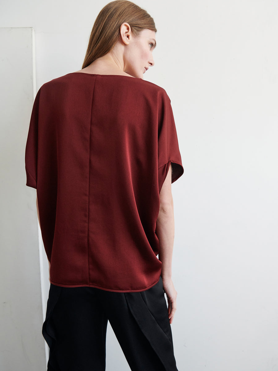 A person with light brown hair stands with their back turned, wearing the loose, short-sleeved Rio Top from Zero + Maria Cornejo in a maroon hue and black pants. They are facing a plain white wall.