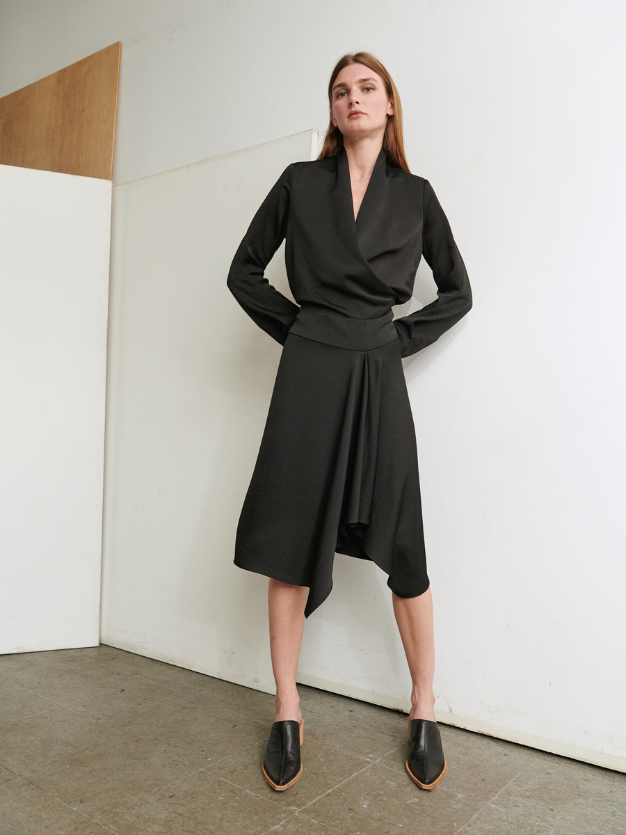 A person with long hair stands confidently with hands on hips, wearing the Long-Sleeved Madira Dress from Zero + Maria Cornejo, a knee-length recycled dress with an asymmetrical hemline. They pair it with black pointed-toe shoes in a minimal indoor space featuring white and wooden panels.