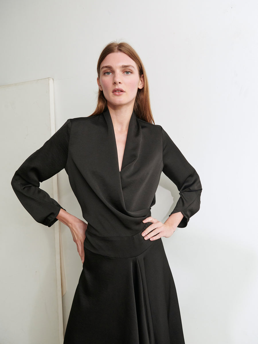 A person with long brown hair is standing against a plain white background. They are wearing the Long-Sleeved Madira Dress by Zero + Maria Cornejo, featuring a draped neckline and an asymmetrical full skirt, exuding elegance. With one hand on their hip, they are looking directly at the camera.