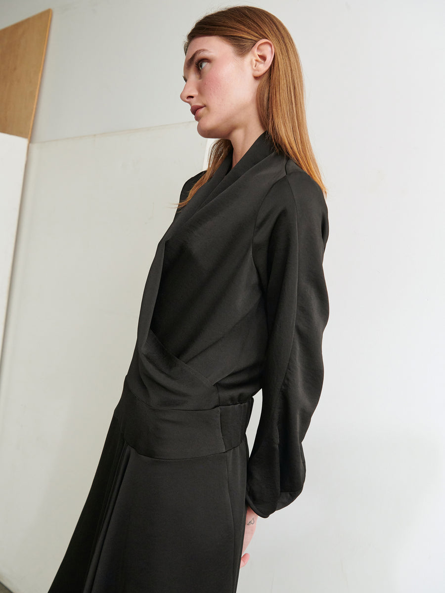 A woman with long, straight hair is standing in profile against a plain, light-colored wall. She is wearing a dark Long-Sleeved Madira Dress by Zero + Maria Cornejo and appears to be looking away from the camera. The background is minimal and simple.