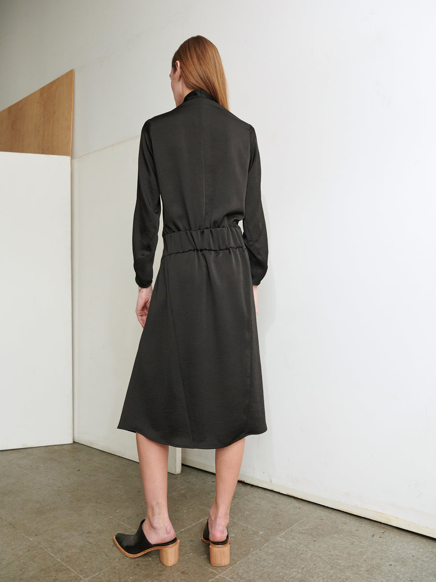 A person with long hair stands facing a white wall in a minimalist room, wearing the elegant Long-Sleeved Madira Dress by Zero + Maria Cornejo. The recycled dress features a knee-length black design with a cinched waist. They are also wearing black open-back shoes with chunky wooden heels. The floor is concrete, and part of a wooden panel is visible.