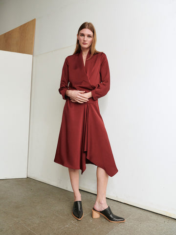 A woman with long brown hair stands indoors, leaning slightly against a wall. She is wearing the Long-Sleeved Madira Dress by Zero + Maria Cornejo in a deep red, paired with black slip-on heels. The background is minimal, featuring plain white walls and a section of wooden paneling.