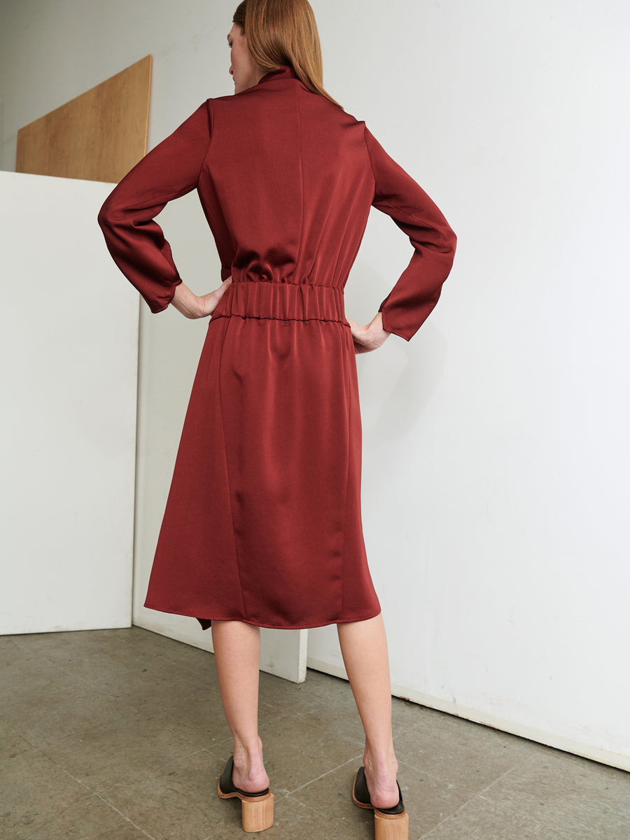 A person stands indoors facing a wall, wearing the Long-Sleeved Madira Dress by Zero + Maria Cornejo. This maroon dress features draped detailing and an asymmetrical skirt that cinches at the waist. The person's back is turned to the camera, and they are sporting open-toed, heeled sandals. The background includes a plain, light-colored wall and a section of wooden paneling.