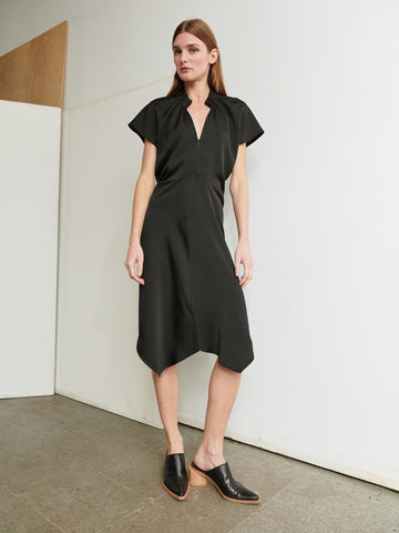 A person with long, straight hair wears the Ruched Stella Dress by Zero + Maria Cornejo, a simple knee-length black dress featuring short sleeves and a front zipper. They are standing next to a white wall with one hand resting by their side and the other slightly bent. They wear black mules with wooden heels and stand on a tiled floor.