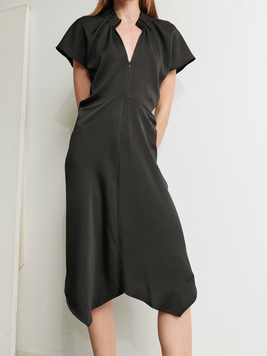 A person wearing the Ruched Stella Dress by Zero + Maria Cornejo stands against a plain, light-colored background. The knee-length, dark dress made from recycled polyester features short sleeves, a V-neck, an asymmetrical hemline, and a seam running down the middle from the neckline to the hem. The person's head and feet are not visible.