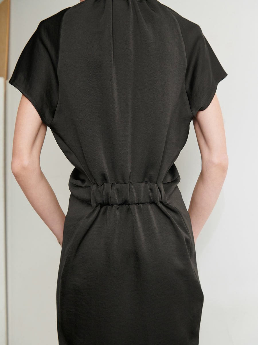 A person is shown from behind wearing the Ruched Stella Dress from Zero + Maria Cornejo, a sleek, black, knee-length dress featuring a gathered waistline. The minimalist design of this recycled polyester dress complements the relaxed posture of their arms at their sides. The background is plain and light-colored.