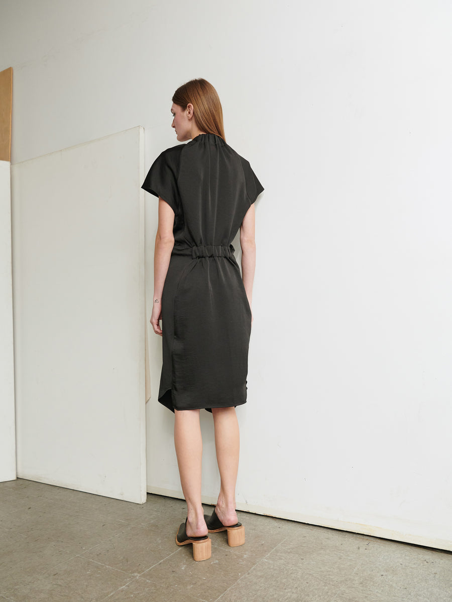 A person with long hair stands facing a wall, clad in the Ruched Stella Dress by Zero + Maria Cornejo, a black recycled polyester dress featuring short sleeves and a belt at the waist. They are wearing open-toed wooden platform shoes and standing on a concrete floor. The minimalistic background consists of white and beige walls.