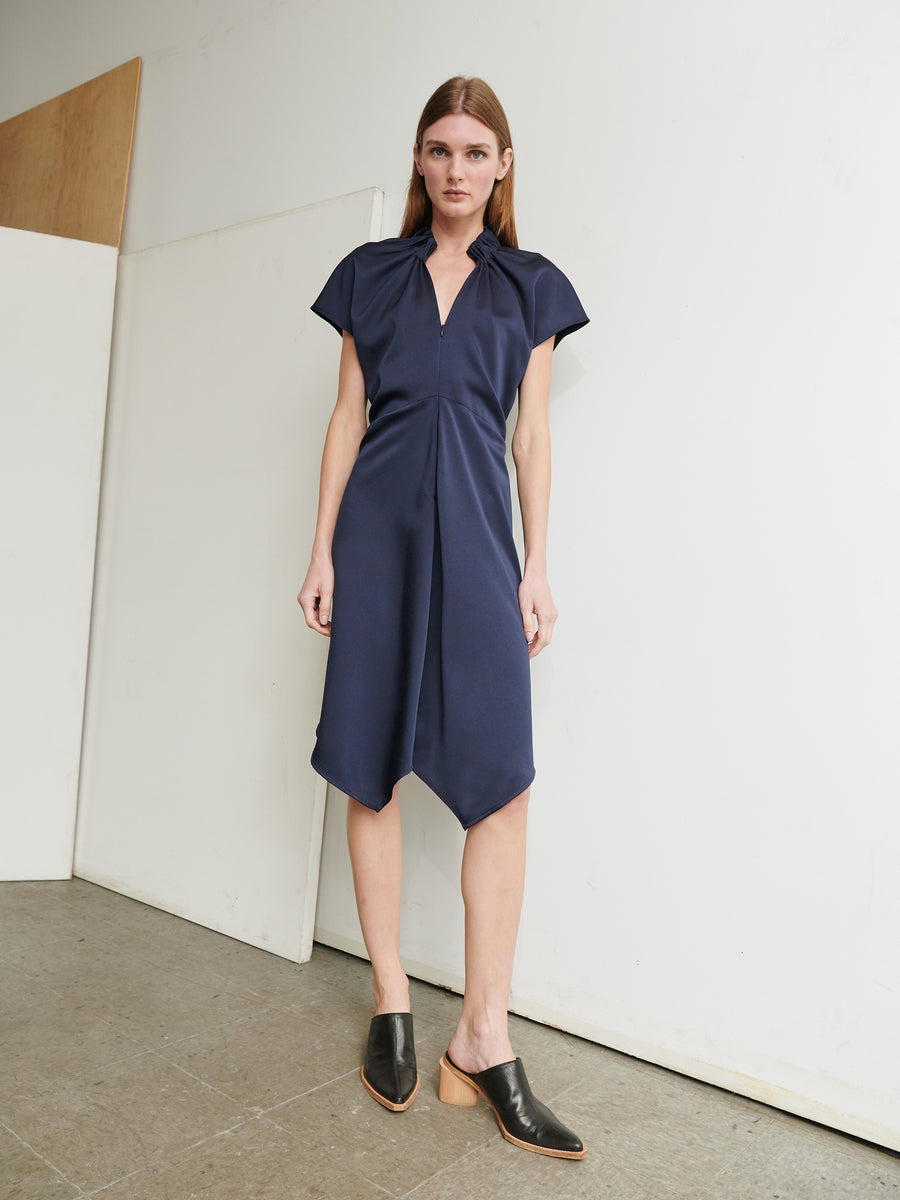 A woman stands in a minimalist indoor setting wearing the Ruched Stella Dress by Zero + Maria Cornejo, which is knee-length, navy blue, with a v-neck and asymmetrical hemline, made from ultra-soft recycled fabric. She pairs the dress with black mules featuring wooden heels. The background is plain with a tiled floor and white wall.