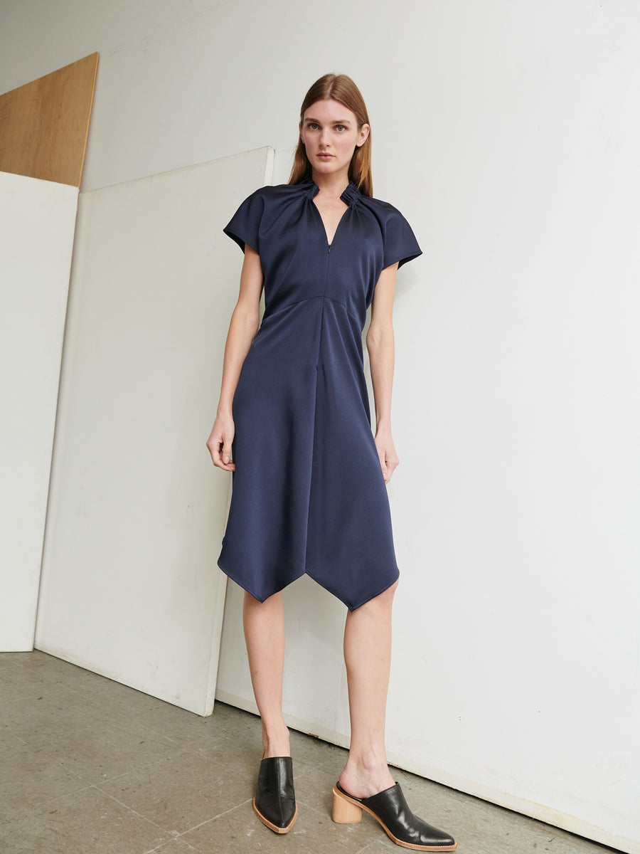 A woman with long brown hair is standing against a white wall and wooden panel, wearing the Ruched Stella Dress in navy blue recycled polyester by Zero + Maria Cornejo. The dress features a V-neck and an asymmetrical hemline. She completes her look with black, heeled mules. The setting appears to be an indoor, minimalistic environment.