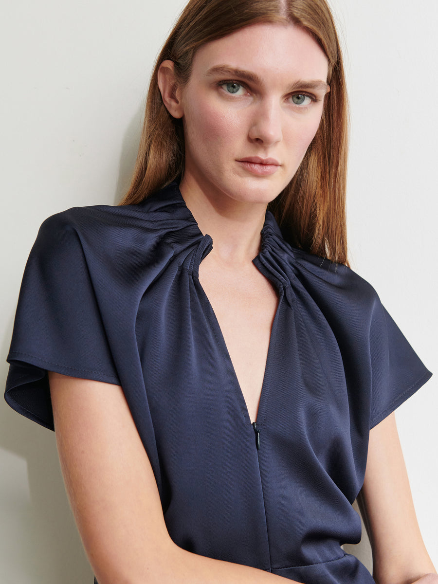 A woman with long brown hair is wearing the Ruched Stella Dress by Zero + Maria Cornejo, a knee-length navy blue dress made from ultra-soft recycled fabric. It features short sleeves and a gathered neckline. She is looking directly at the camera against a plain, light-colored background.