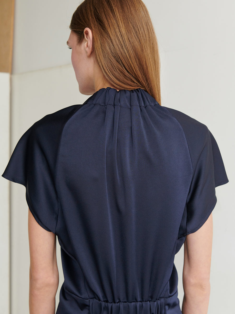 A person with long, light brown hair is seen from the back, wearing the Ruched Stella Dress by Zero + Maria Cornejo, a dark navy blue garment made from ultra-soft recycled fabric with a gathered neckline and short sleeves. The wall behind them is light grey and beige.