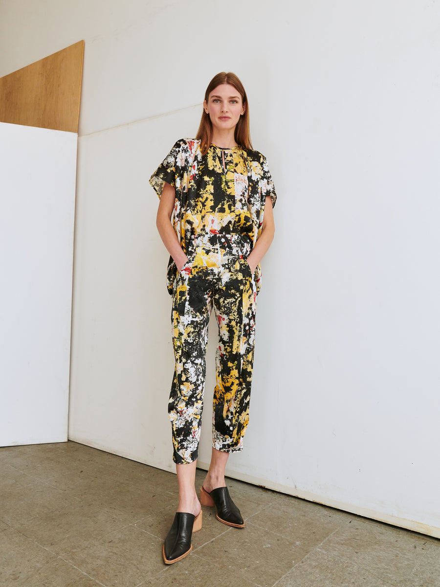 A woman stands indoors against a white wall with a wooden panel. She is wearing the Cropped Akeo Pant from Zero + Maria Cornejo, paired with a matching floral print top made from stretch silk charmeuse. The outfit features short sleeves and cropped pants in black, yellow, and white. Her hands are in her pockets, and she completes the look with black pointed-toe mules.