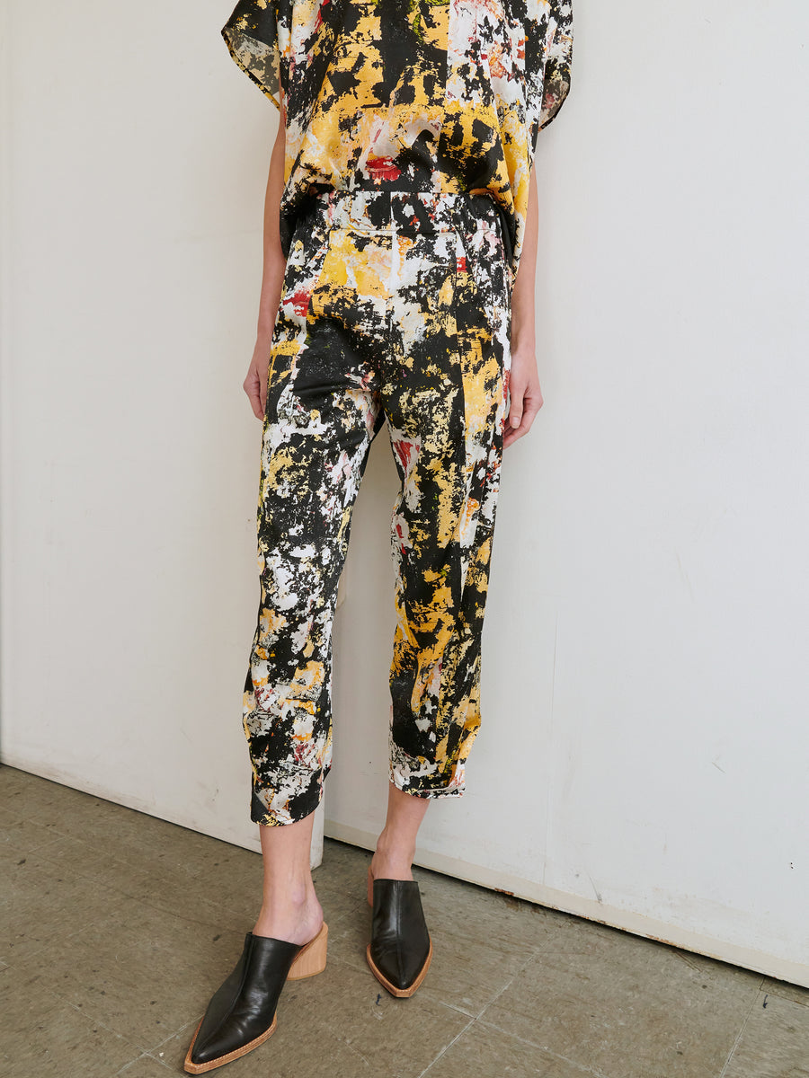 A person standing against a plain wall is wearing a matching stretch silk charmeuse outfit featuring a vibrant abstract yellow, black, and white pattern. Complementing the look are the Cropped Akeo Pant by Zero + Maria Cornejo and black mules with wooden heels. The image captures their outfit from the neck down.