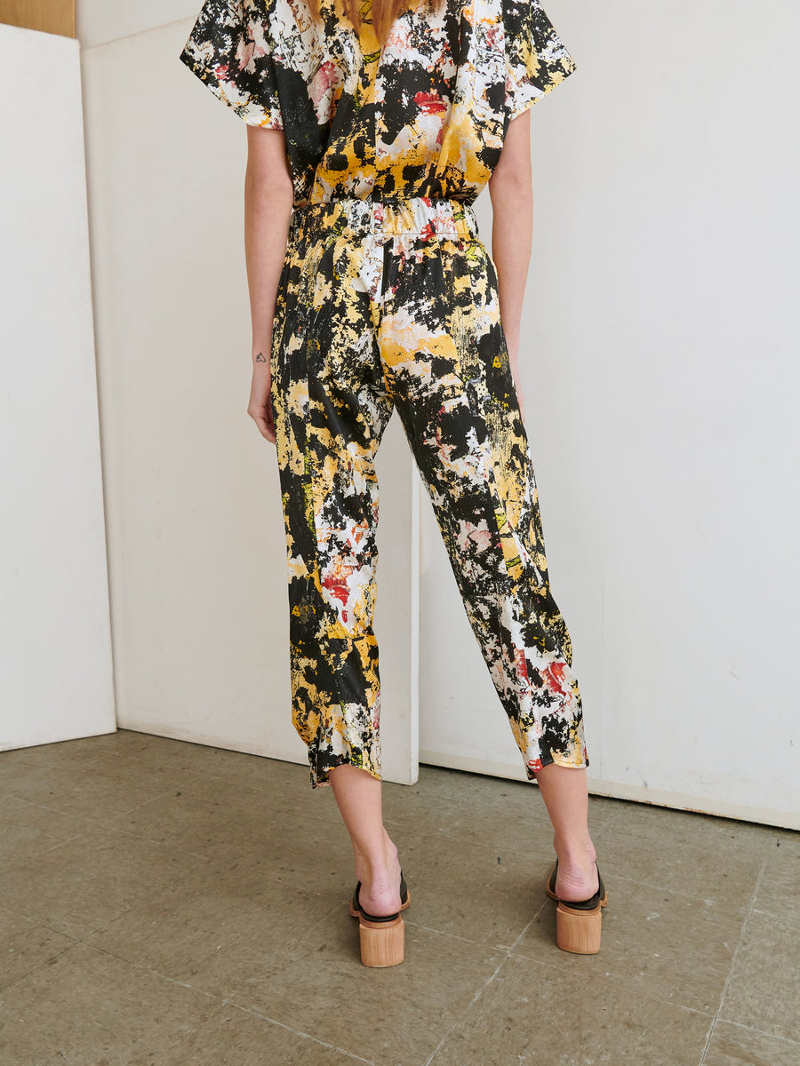 A person stands with their back to the camera, dressed in a colorful, abstract-print Cropped Akeo Pant made of stretch silk charmeuse from Zero + Maria Cornejo. They are wearing platform sandals with wooden block heels. The background features white and beige walls and grey flooring.