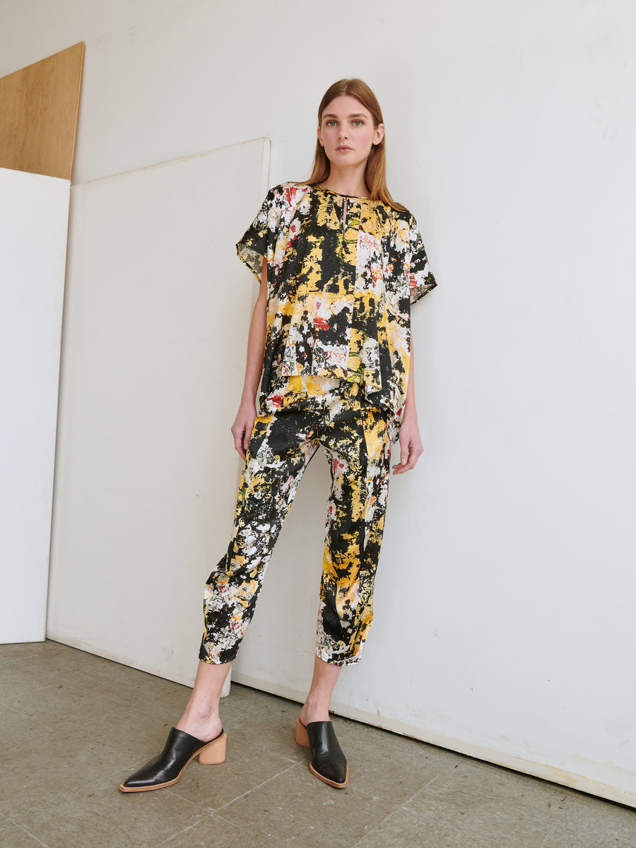 A woman stands against a plain wall wearing the Rae Poncho Top from Zero + Maria Cornejo, paired with matching cropped pants featuring a vibrant abstract pattern in yellow, black, white, and red. She has long hair and wears black slip-on shoes with wooden block heels.