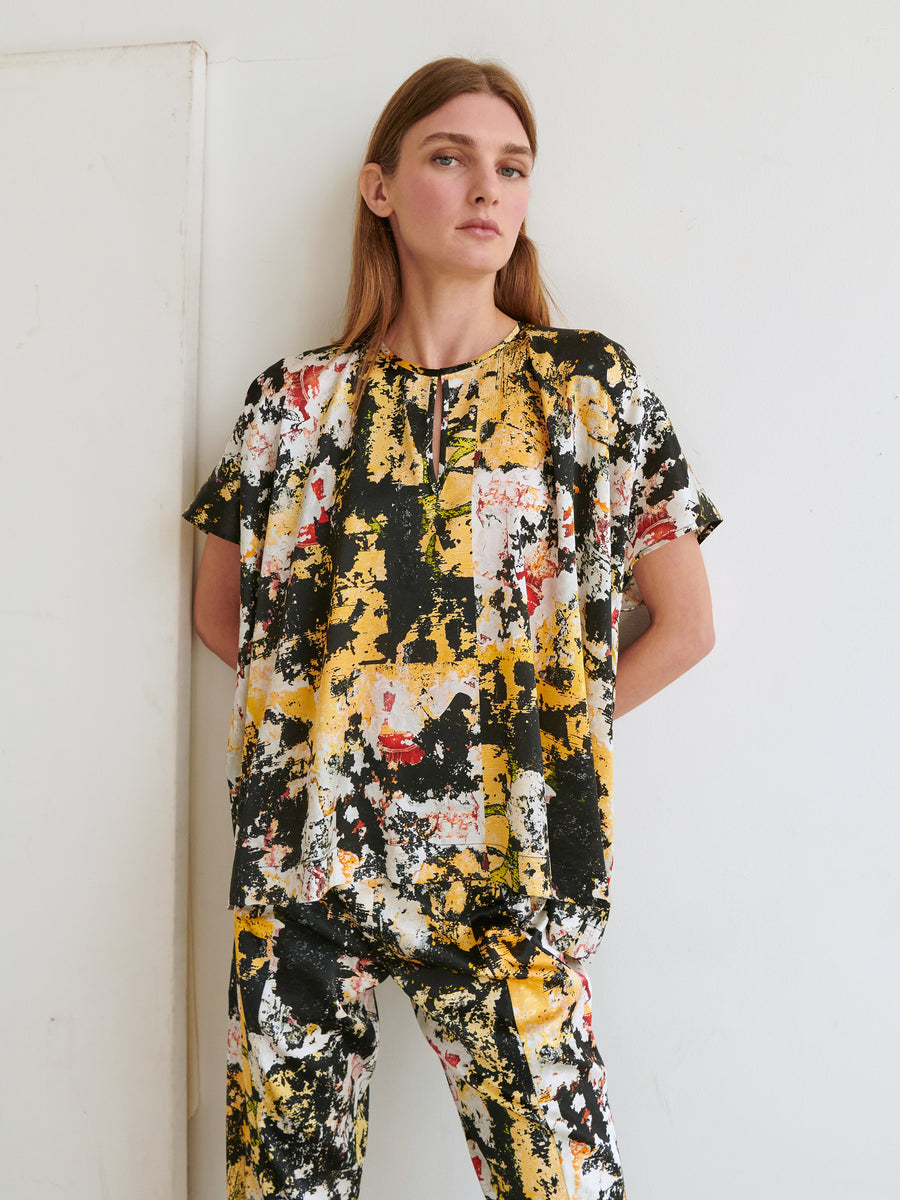 A woman with long, straight hair is standing against a white wall, wearing the Rae Poncho Top from Zero + Maria Cornejo along with matching pants. The set is made of stretch silk charmeuse and features an abstract, colorful pattern with black, yellow, white, and red splashes. She looks directly at the camera with a neutral expression.