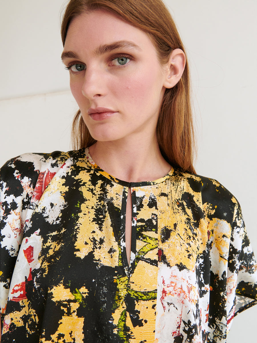 A woman with long, straight brown hair is wearing the Rae Poncho Top by Zero + Maria Cornejo, made of stretch silk charmeuse. The top showcases a colorful, abstract print in shades of yellow, black, white, and red with a front keyhole cutout. She is looking at the camera with a neutral expression against a plain, light-colored background.