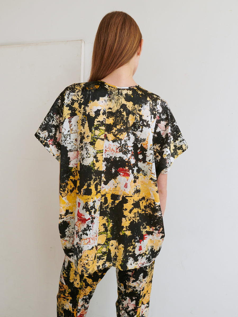 A person with long hair, seen from behind, is wearing the Rae Poncho Top by Zero + Maria Cornejo, along with a coordinating set of loose-fitting pants. The ensemble features an abstract print in black, yellow, white, and red hues. The background is a plain white wall.