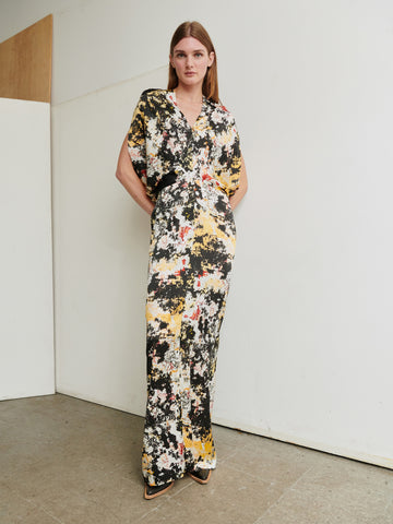 A person with long hair stands against a plain off-white wall, wearing the Long Reni Dress from Zero + Maria Cornejo. The floor-length dress features an abstract floral print in shades of black, yellow, and white. It has wide short sleeves, a v-neckline, and they have their hands in the dress's pockets while looking at the camera.