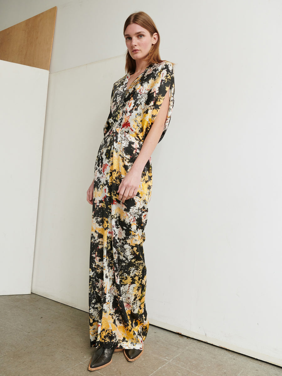 A woman stands in a minimalist room wearing the Long Reni Dress by Zero + Maria Cornejo. The dress, made of stretch silk charmeuse, features yellow, black, and white splashes of color in a graphic print. She pairs it with black pointed shoes and has straight brown hair, looking confidently at the camera.