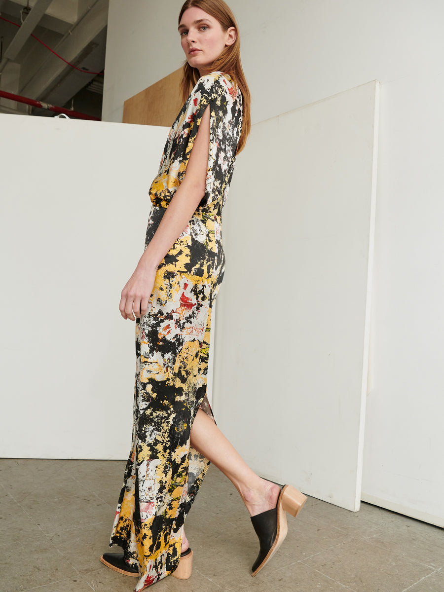 A woman with long hair is standing sideways in an indoor setting, wearing the Long Reni Dress by Zero + Maria Cornejo. The dress is made of stretch silk charmeuse with an abstract print. She pairs it with black shoes that feature a wooden heel. The minimalistic background consists of white panels and a concrete floor.