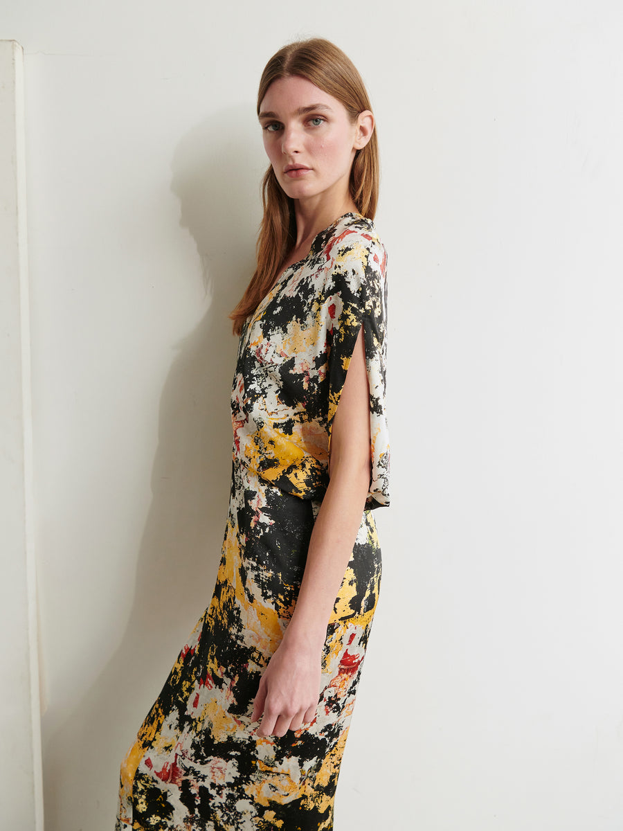 A person with long hair stands against a white wall, wearing the Long Reni Dress by Zero + Maria Cornejo, which features a vibrant splattered paint design in black, yellow, white, and red. They look directly at the camera with a calm expression, their right hand resting by their side.