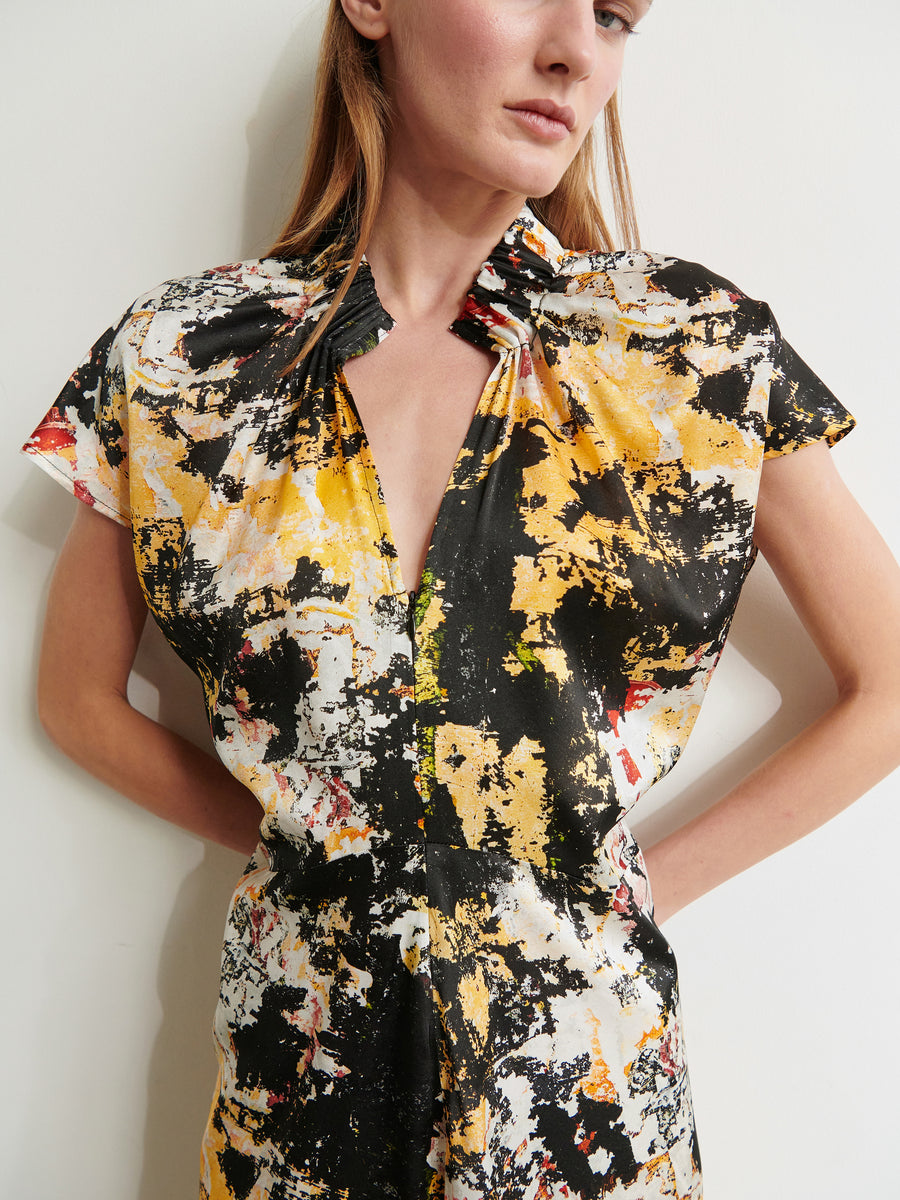 A person stands against a plain backdrop wearing the Ruched Stella Dress by Zero + Maria Cornejo. The short-sleeved dress features an abstract, digital inkjet paint-splatter pattern in black, white, yellow, and hints of red. It has a V-neck and gathered details at the shoulders and chest. The person has long, light brown hair.