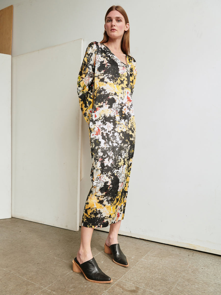 A person stands against a plain background wearing the Long-Sleeved Off Kilter Dress from Zero + Maria Cornejo, featuring an abstract pattern in shades of yellow, black, and white. They are also wearing black mules with wooden heels. The person has long hair and stands with hands in their pockets, looking forward.