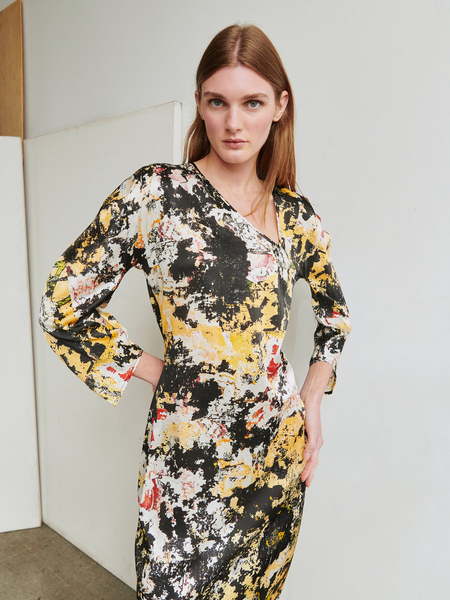 A woman with straight, light brown hair stands against a plain background. She is wearing the Long-Sleeved Off Kilter Dress from Zero + Maria Cornejo, made from stretch silk charmeuse and featuring an abstract pattern with splashes of yellow, black, white, and red. Her asymmetrical v-neckline adds a touch of elegance as she gazes directly at the camera.