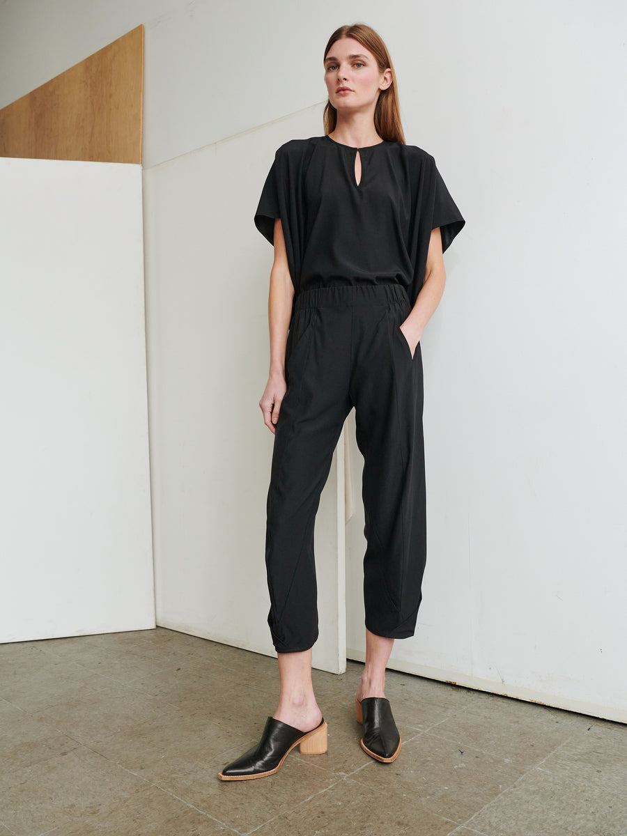 A woman stands against a minimalist indoor background wearing Zero + Maria Cornejo’s Cropped Akeo Pant, a black, loose-fitting jumpsuit with short sleeves, an elastic waist, and a keyhole neckline. She poses with hands in her pockets, accentuating the cropped pant style and pairs it effortlessly with black heeled mules.