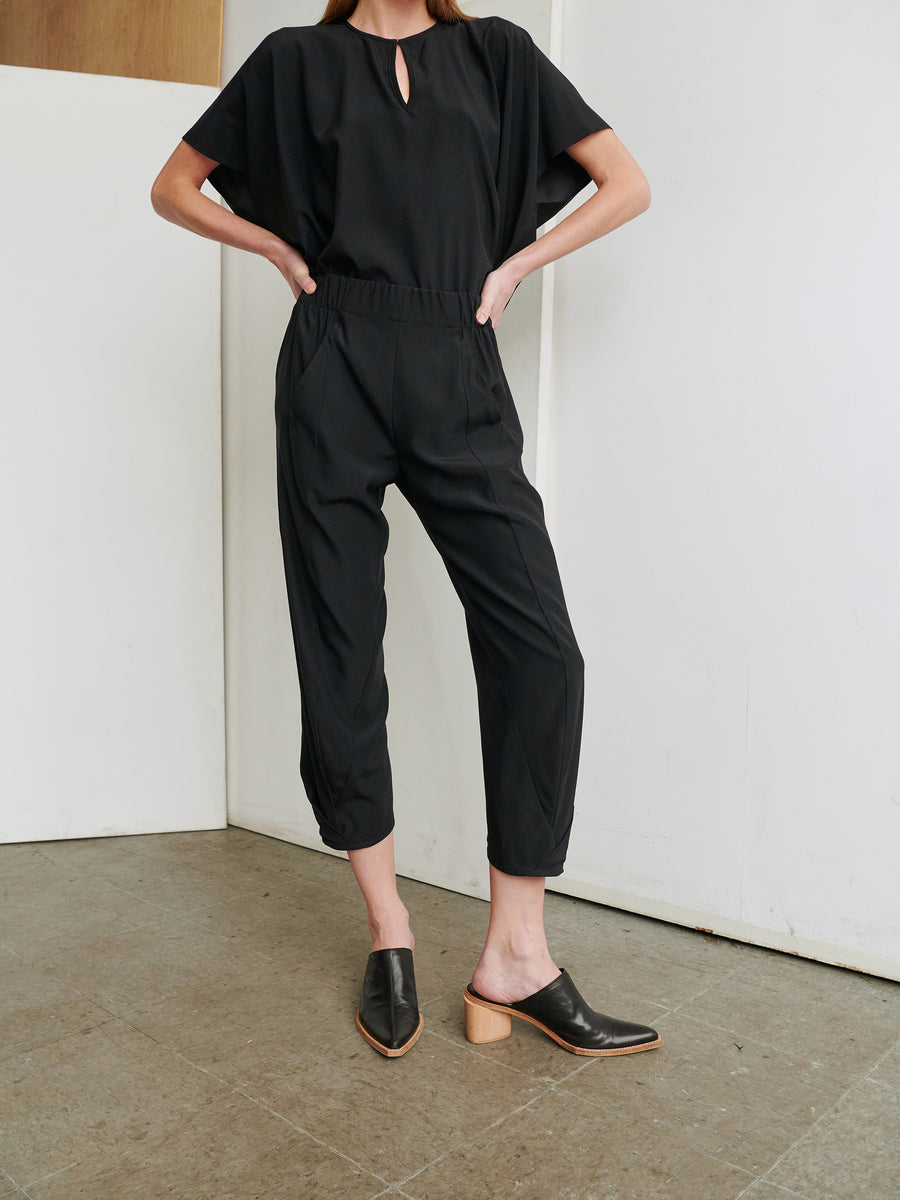 A person stands against a plain wall, wearing the Cropped Akeo Pant by Zero + Maria Cornejo. The black jumpsuit features short, wide sleeves and cropped pant legs made of silky fabric reminiscent of stretch silk charmeuse. They have their hands tucked into the pockets and complement the outfit with black mule shoes that have wooden heels.